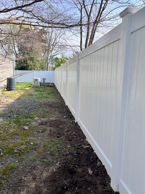 White vinyl fence along a narrow dirt path beside a yard with trees and a small shed