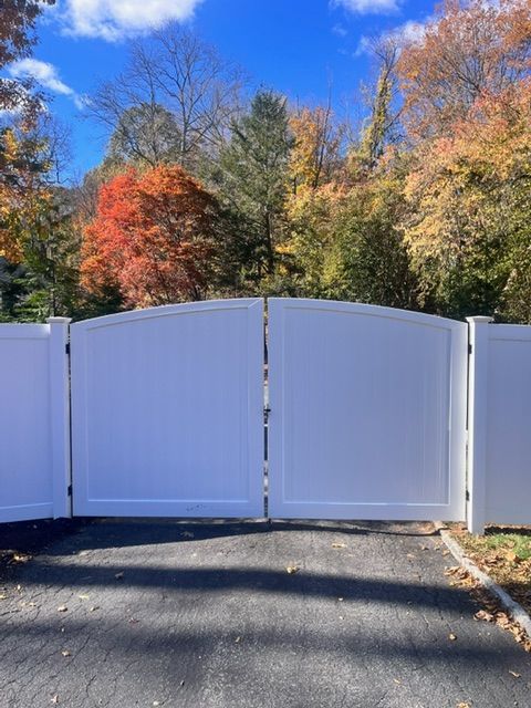 White, arched driveway gate with asphalt in front and colorful autumn trees in the background.