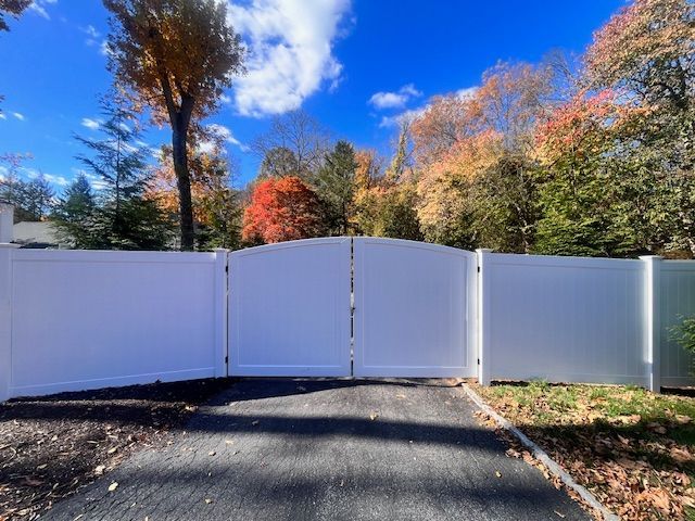 White vinyl fence with double gate on a paved driveway; autumn trees with colorful leaves in the background.
