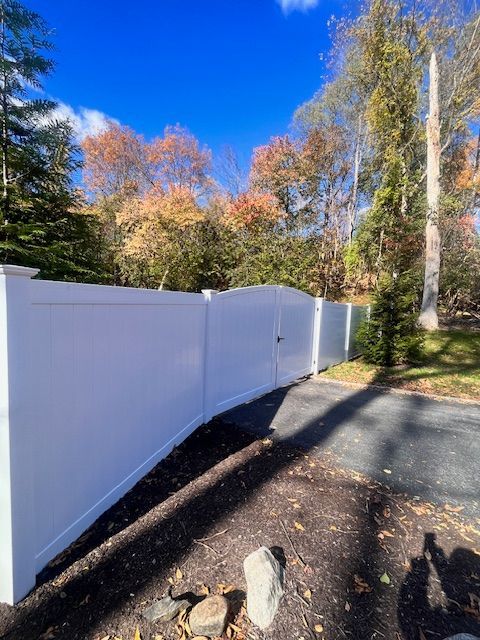 White vinyl fence with gate, bordering a driveway and yard with trees under a blue sky.