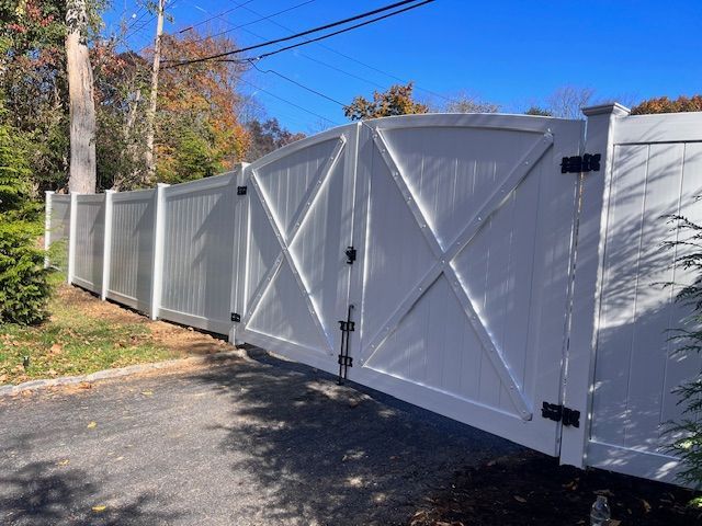 White wooden gate with an X design, flanked by a white fence on a paved driveway in a yard.