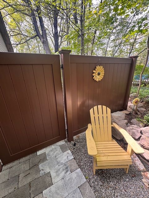 Wooden Adirondack chair beside a brown fence and gate in a gravel patio with trees behind