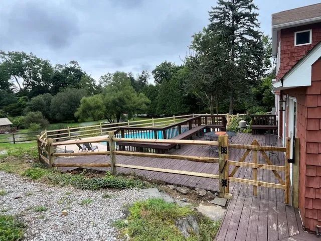 Wooden deck with a pool enclosed by a wooden fence, next to a red-sided building and a grassy area under an overcast sky.