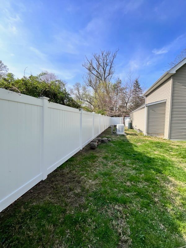 Narrow grassy side yard with white vinyl fence, beige house, and blue sky