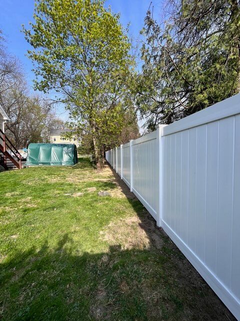 Backyard with green lawn, trees, and a long white privacy fence on the right