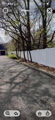 Tree-lined driveway beside a white fence and small building on a sunny day