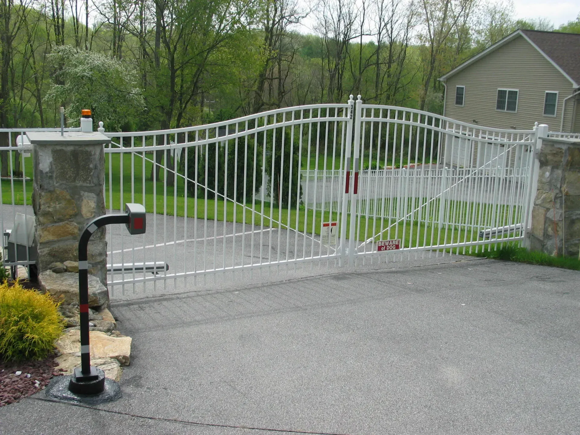 White security gate on a paved driveway, leading to a house in a green yard. Gate is between stone pillars.