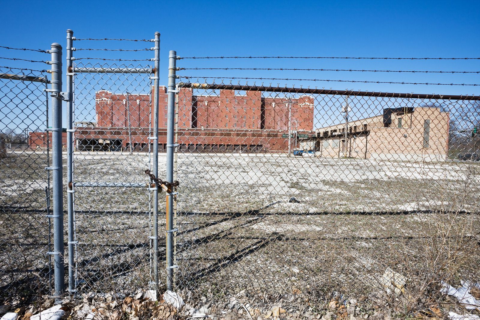 A chain-link fence surrounds a brick industrial building on a field with patchy snow and dry grass under a clear blue sky.