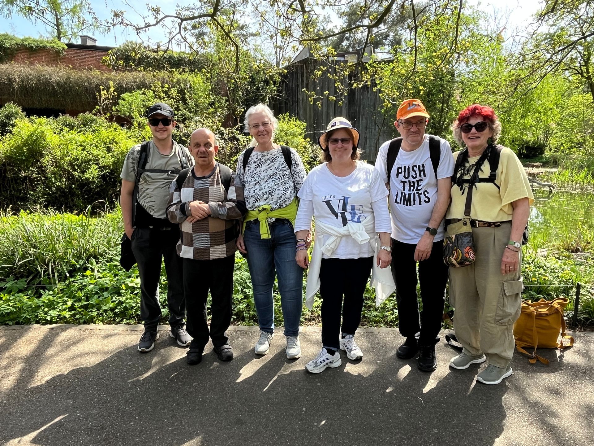 Eine Gruppe von Menschen, darunter einige im Rollstuhl, posiert auf einem Kopfsteinpflasterweg vor einem Holzgebäude mit Torbogen.