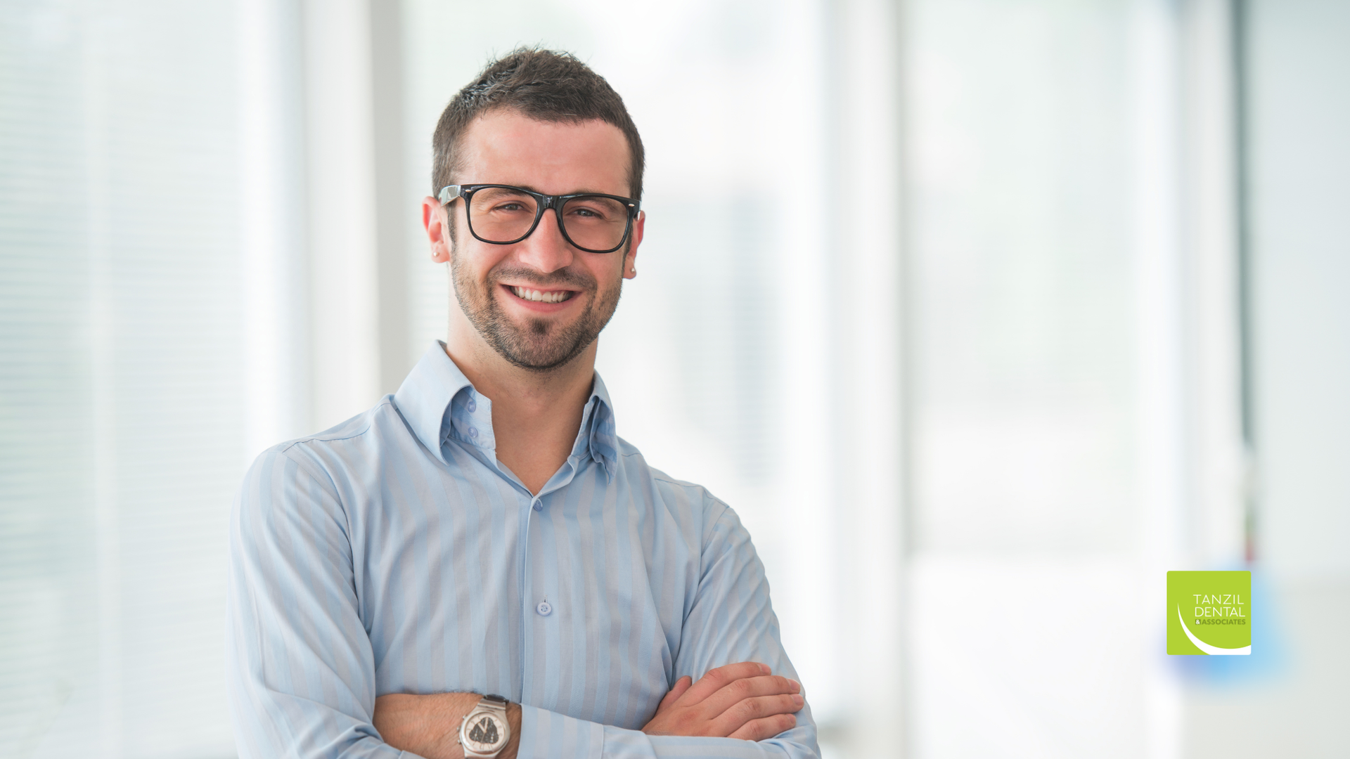 Man wearing glasses smiles, arms crossed, in a brightly lit office setting.