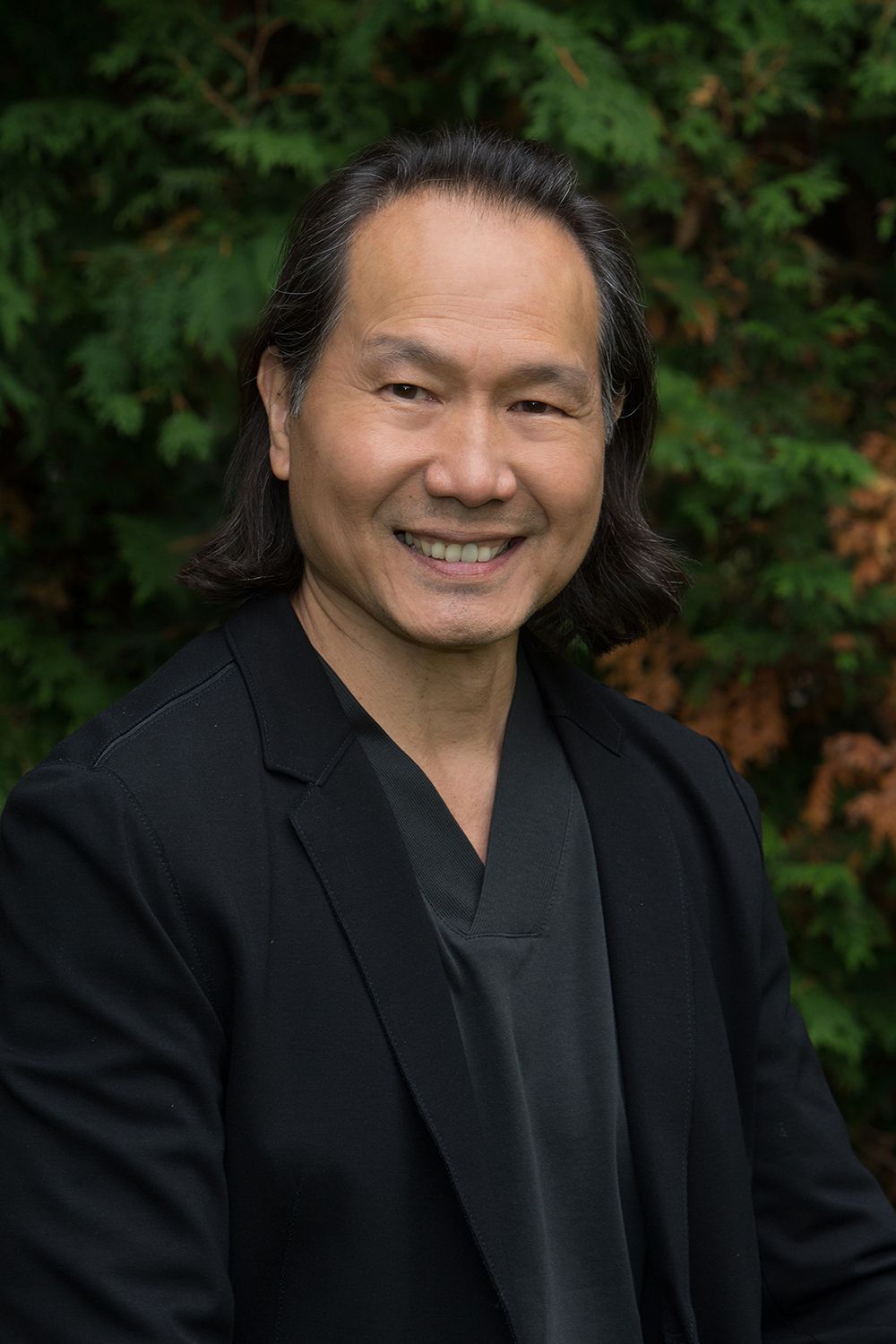 A man with long hair is wearing a black jacket and smiling for the camera.