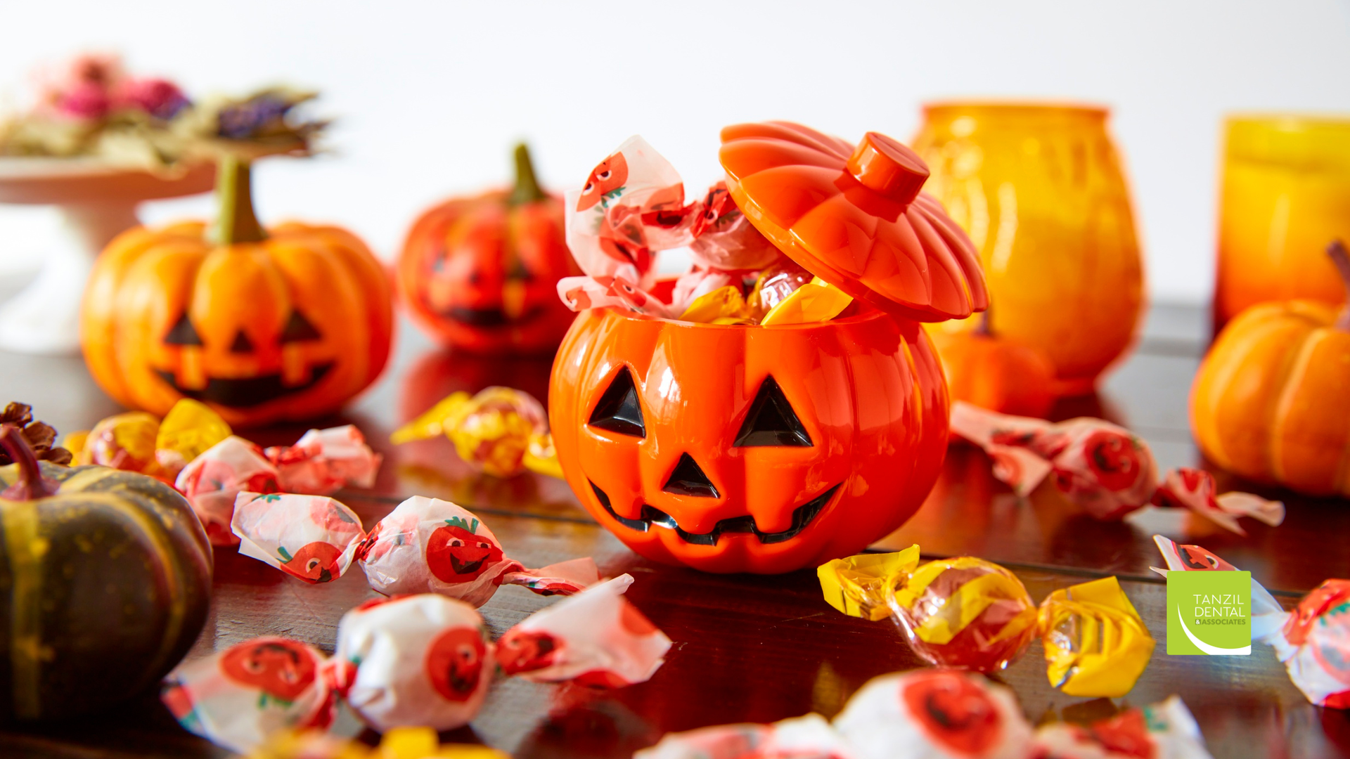 Halloween display: Jack-o'-lantern candy dish overflowing with wrapped candies, surrounded by pumpkins and orange jars.
