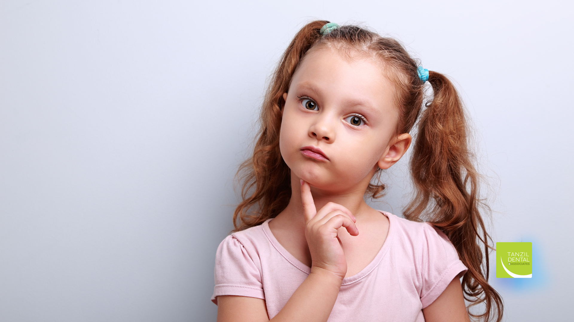 Girl with pigtails holding chin, looking thoughtful, light pink shirt, light blue background.