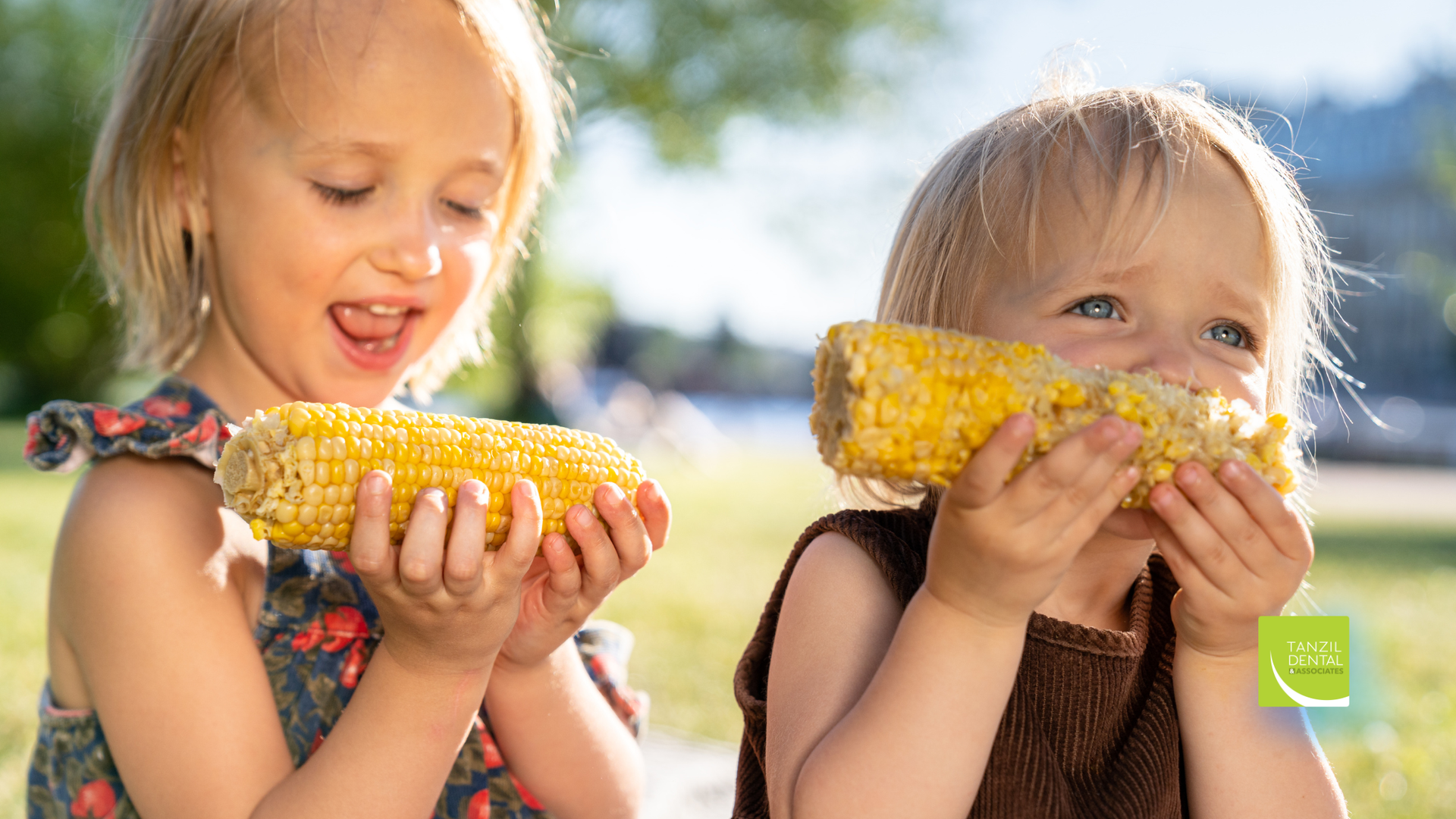 Two children eating corn on the cob outdoors; one smiles, the other looks up.