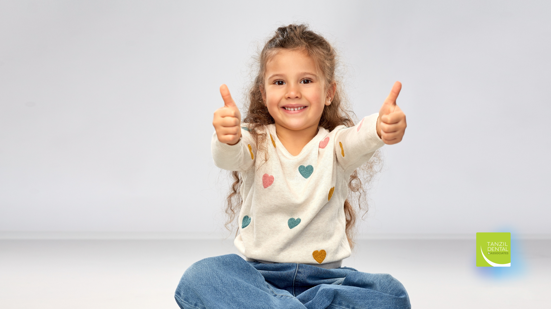 Young girl with curly hair sitting cross-legged, smiling and giving thumbs up against a grey backdrop.