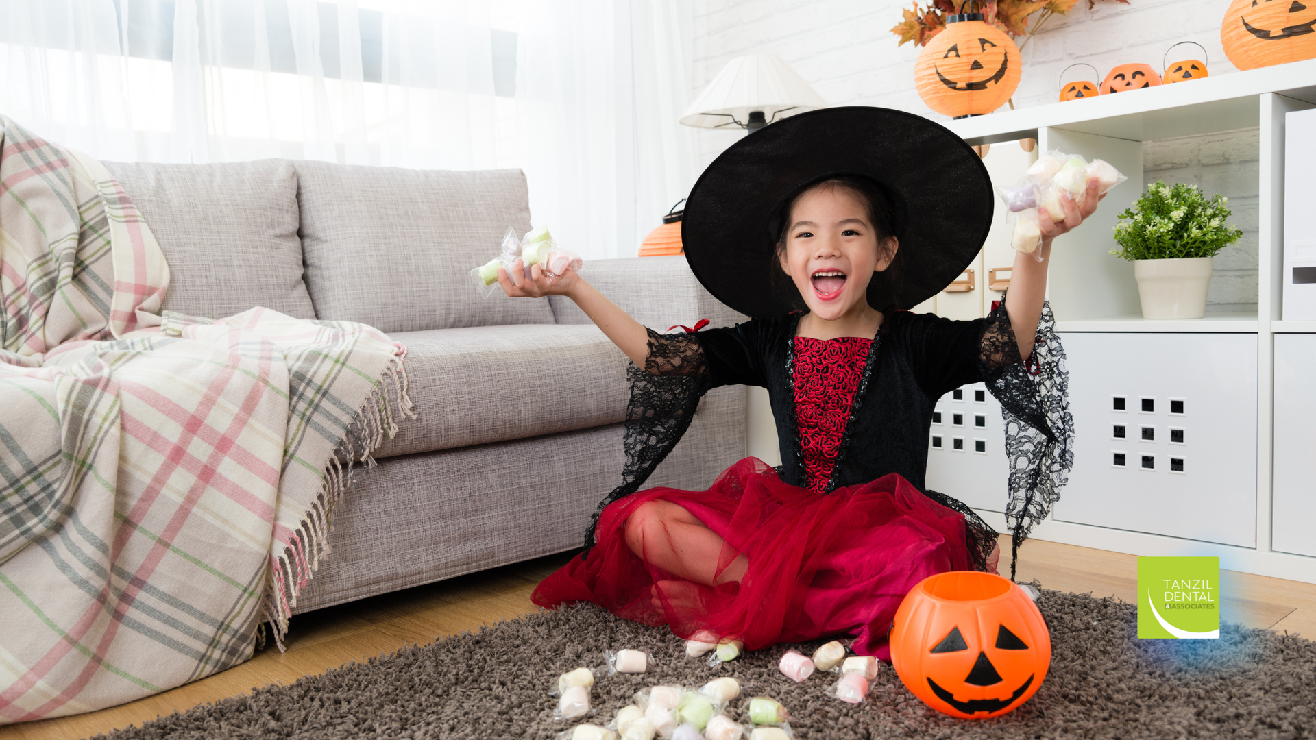 A young girl in a witch costume with arms raised, surrounded by marshmallows and a pumpkin bucket in a decorated room.