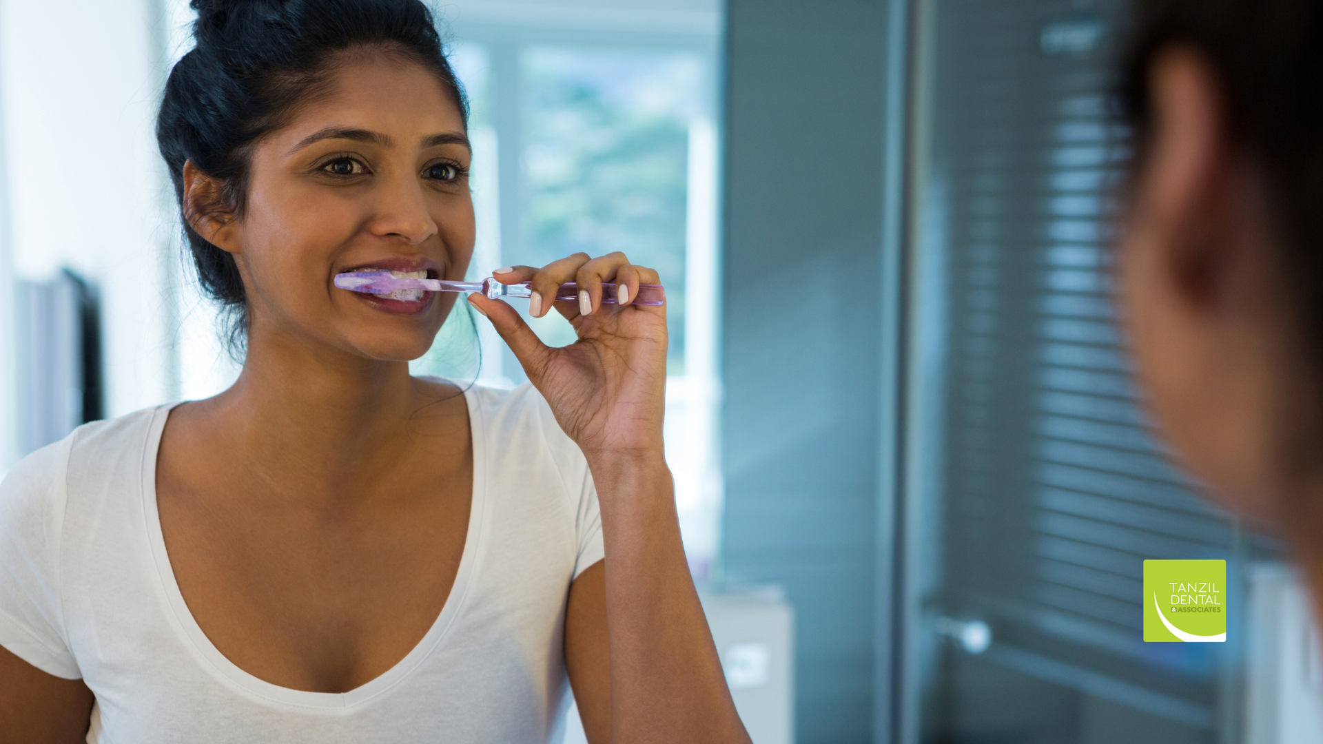 Woman brushing teeth in front of a mirror, holding a toothbrush, in a bathroom.