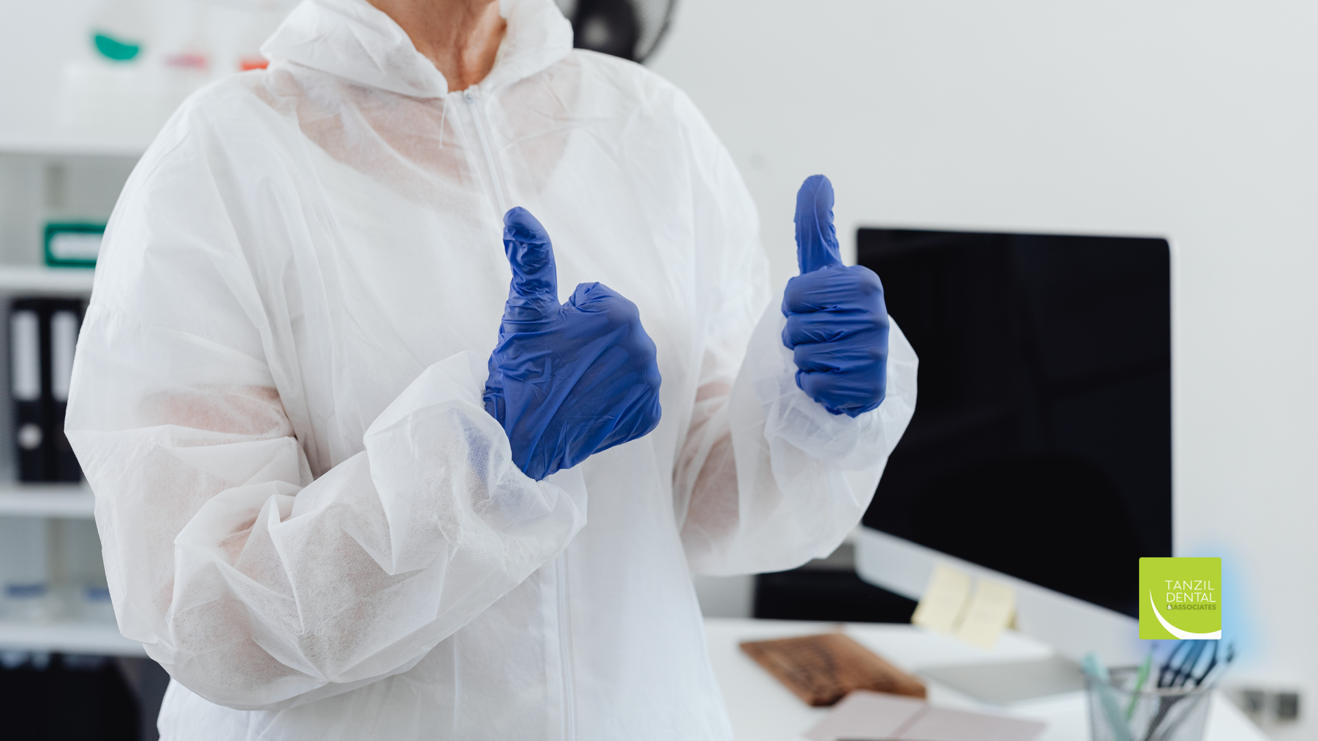 Person in a white protective suit and blue gloves gives a double thumbs-up in an office setting.