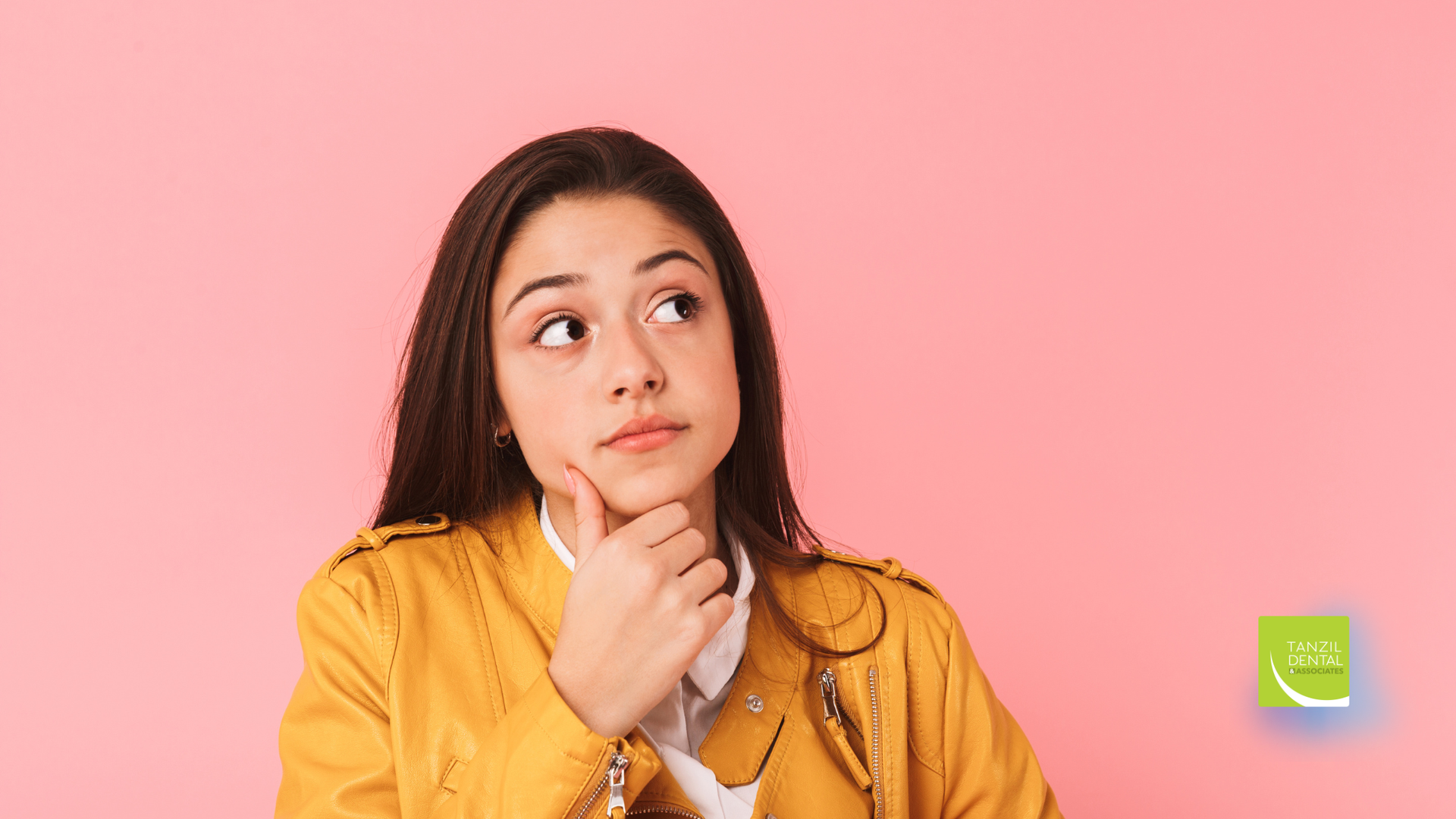 Young woman in yellow jacket, looking up thoughtfully, hand on chin, pink background.