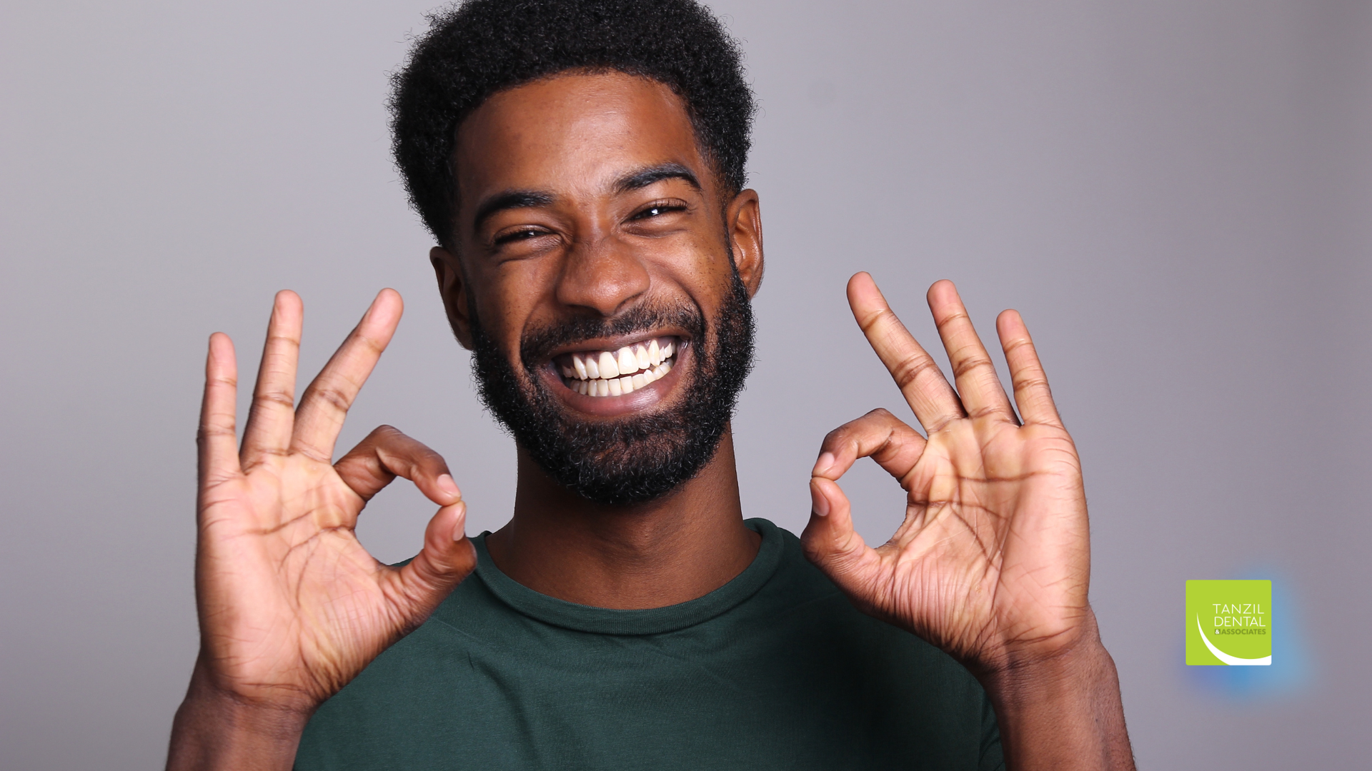 Man with a beard smiling, holding both hands up with “OK” signs. Gray background.