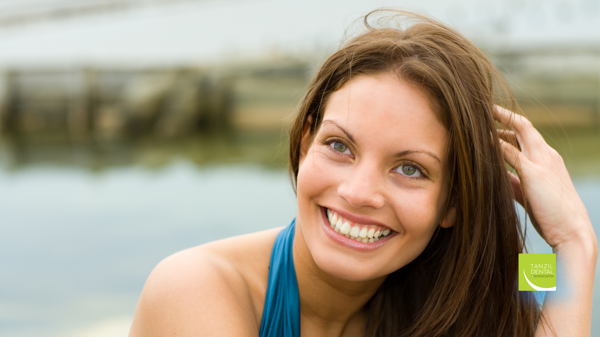 A woman is smiling while holding her hair in front of a body of water.