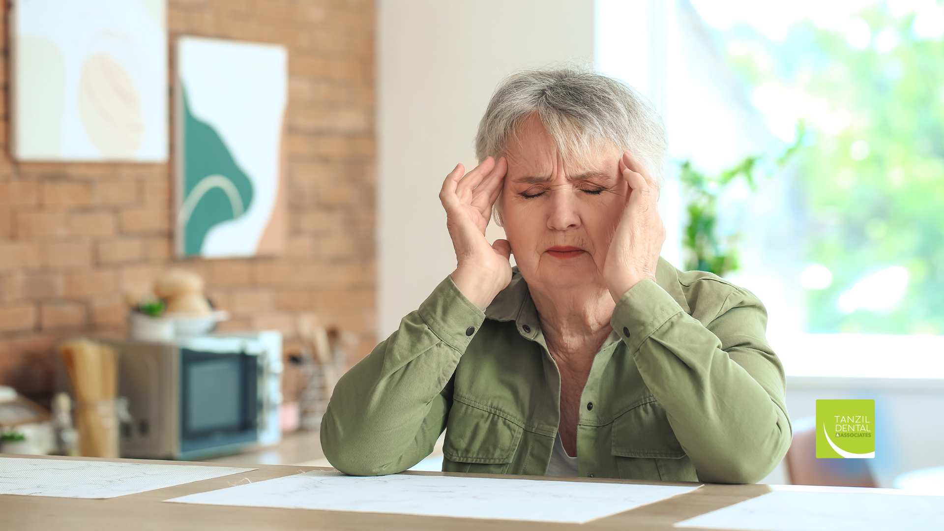 An older woman is sitting at a table with her hands on her head.