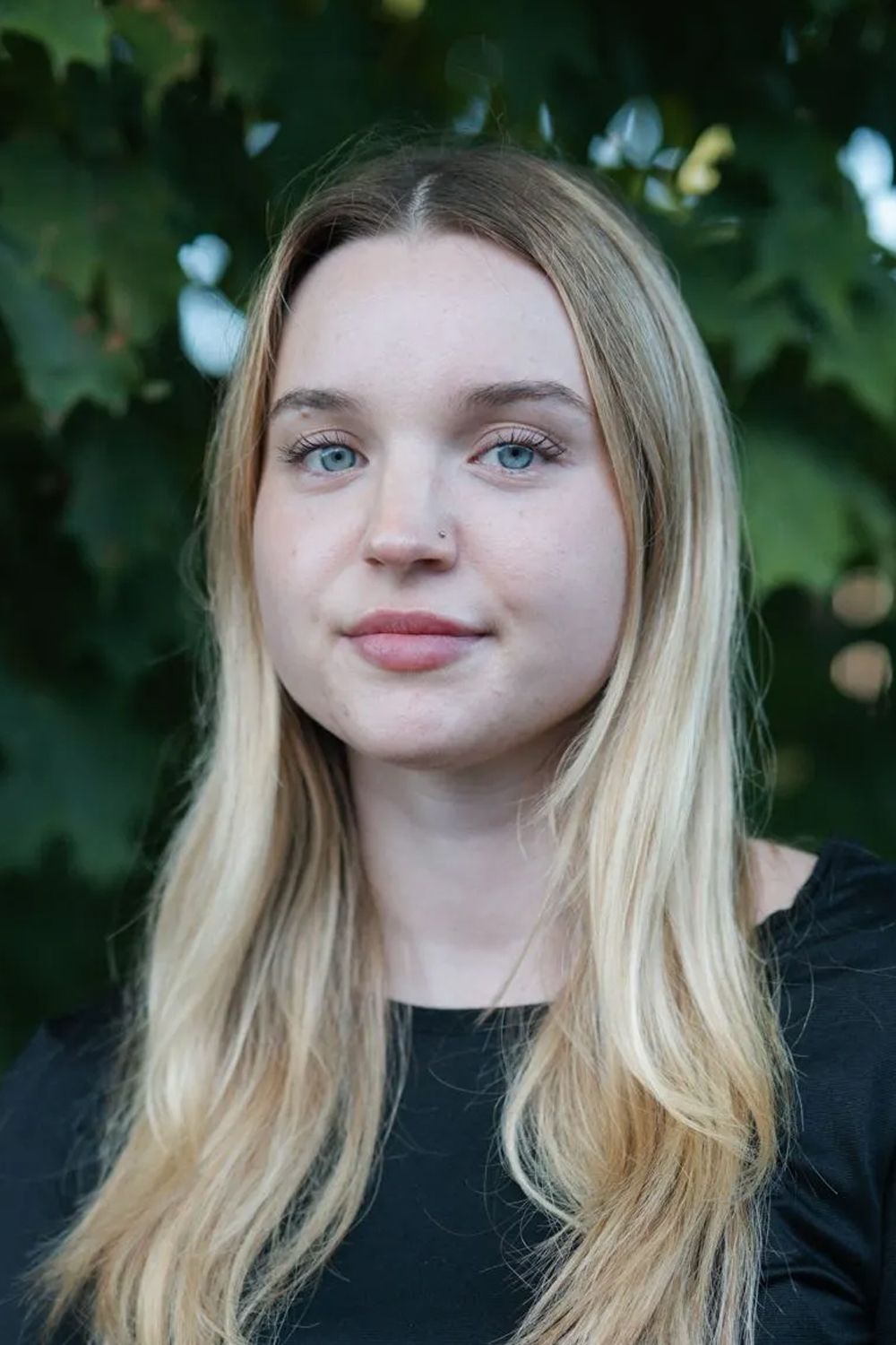 A young woman with long blonde hair and blue eyes is standing in front of a tree.