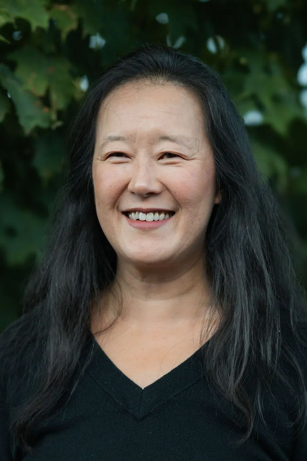 A woman with long black hair is smiling in front of a tree.