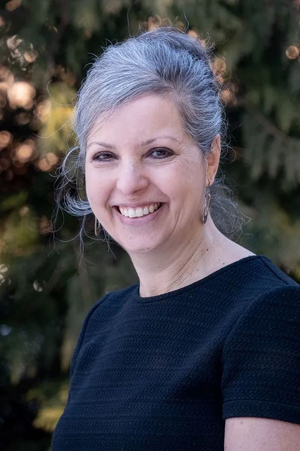 A woman with gray hair is smiling in front of a tree.