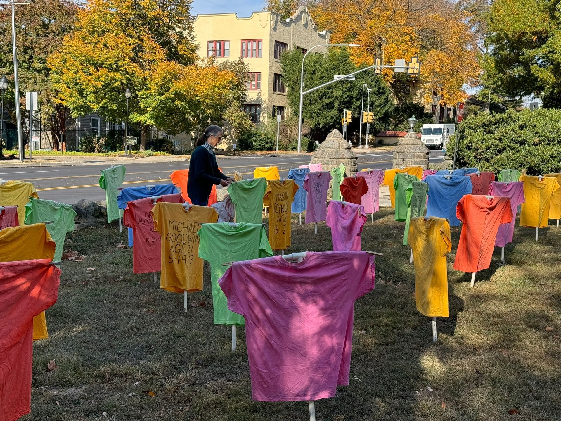 A person arranges colorful t-shirts mounted on stakes in a park-like lawn, creating a vibrant outdoor installation.