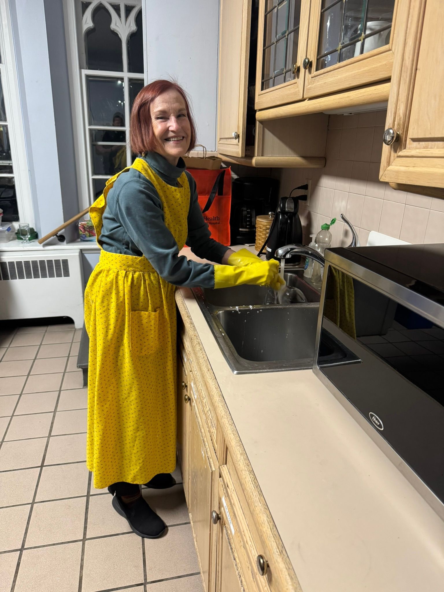 A person wearing a yellow apron and yellow rubber gloves smiles while washing dishes at a kitchen sink.