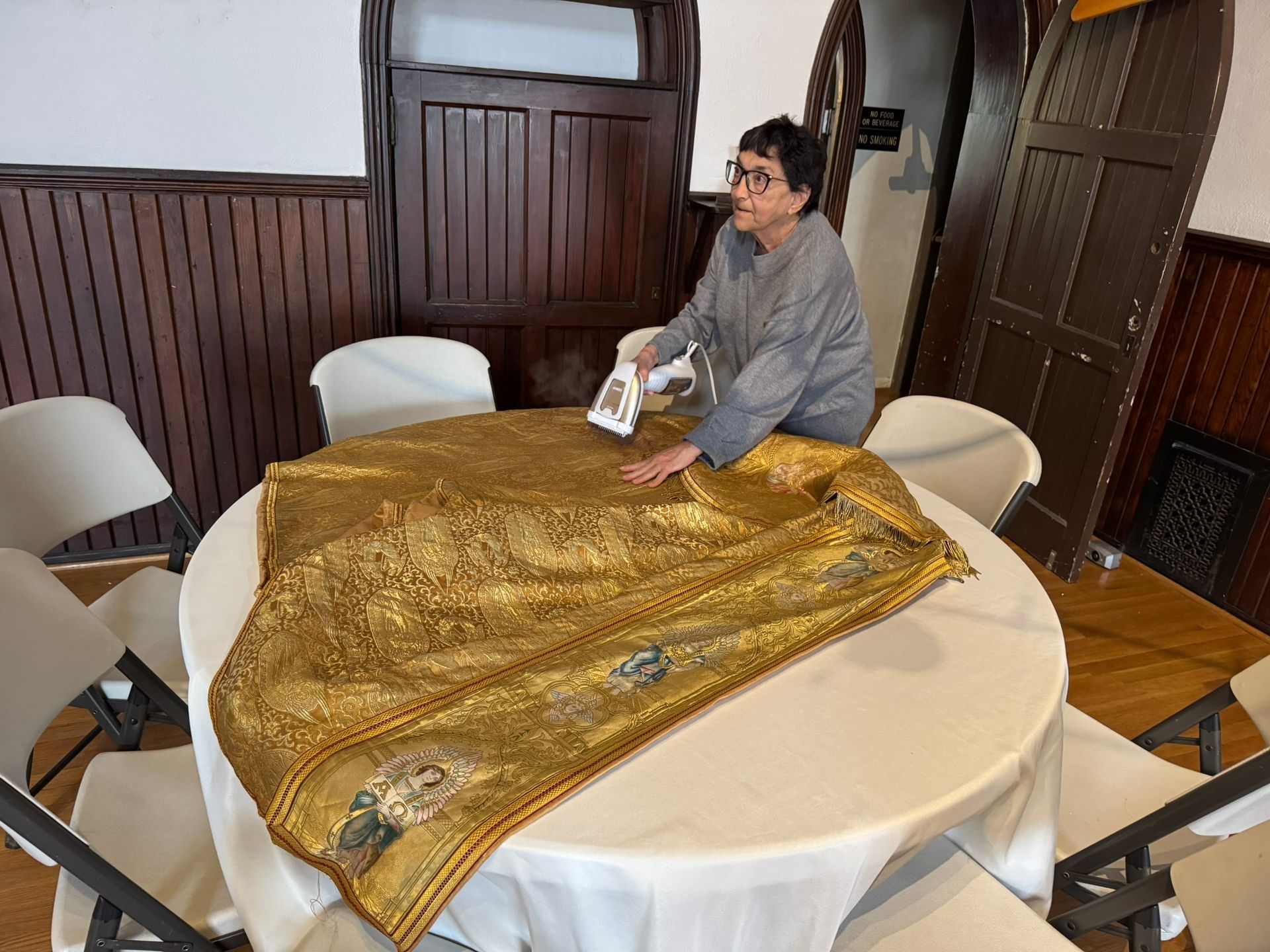 A person using a handheld steamer on a decorative gold fabric laid over a round white table in a room with wood paneling.