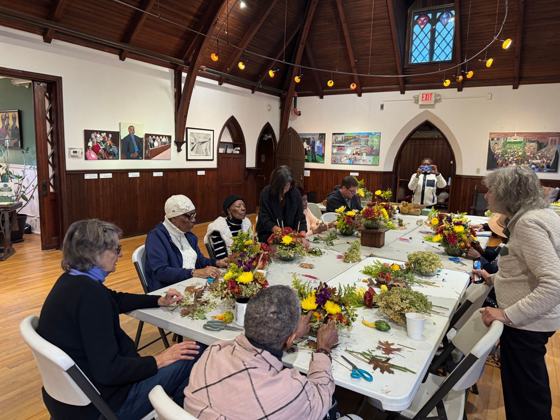A group of people sit around tables in a hall, collaboratively arranging floral centerpieces with fall-themed foliage.