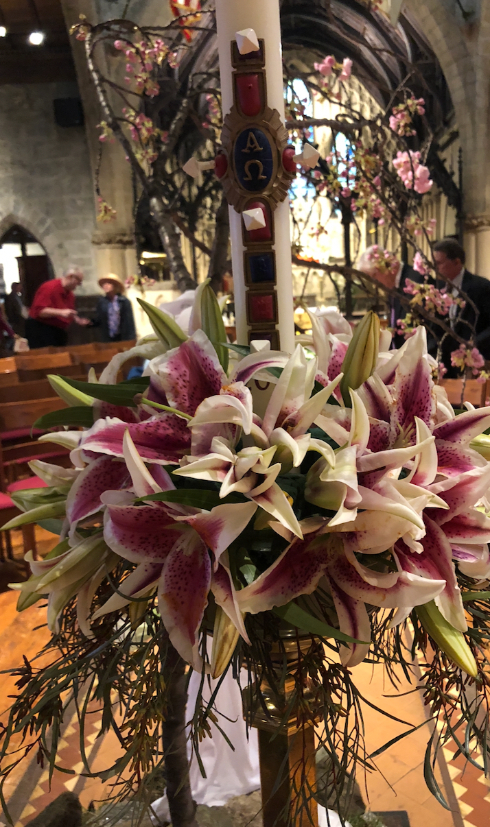 A Paschal candle with an Alpha and Omega emblem stands behind a large bouquet of pink and white lilies in a church.