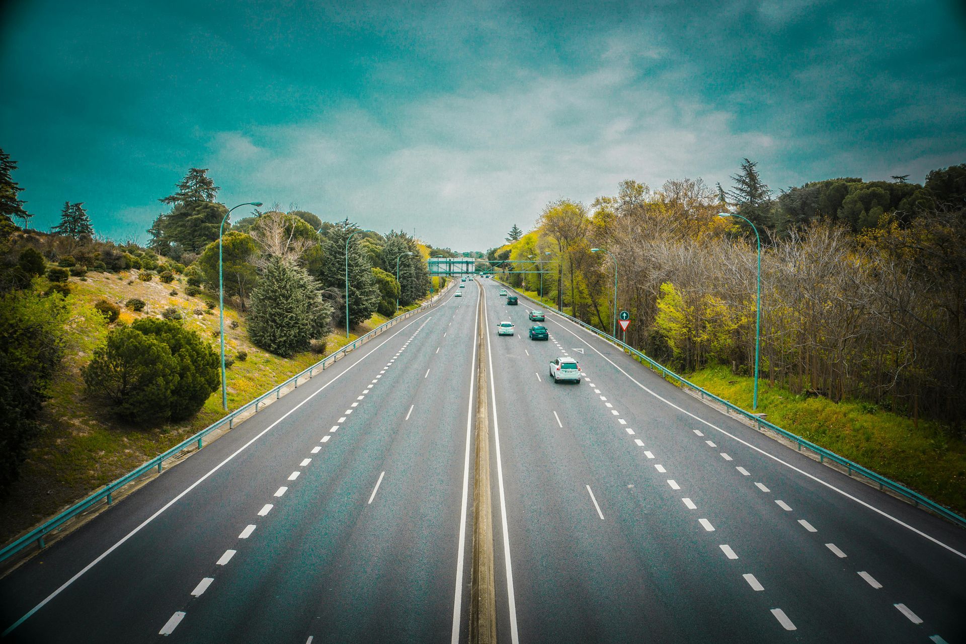 Highway with cars driving under a cloudy sky, trees line both sides of the road.