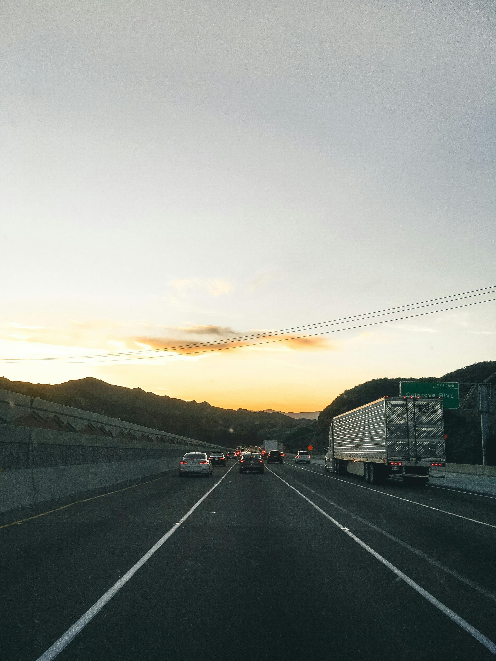 Highway at sunset with cars and a semi-truck; mountains on both sides, birds flying overhead.