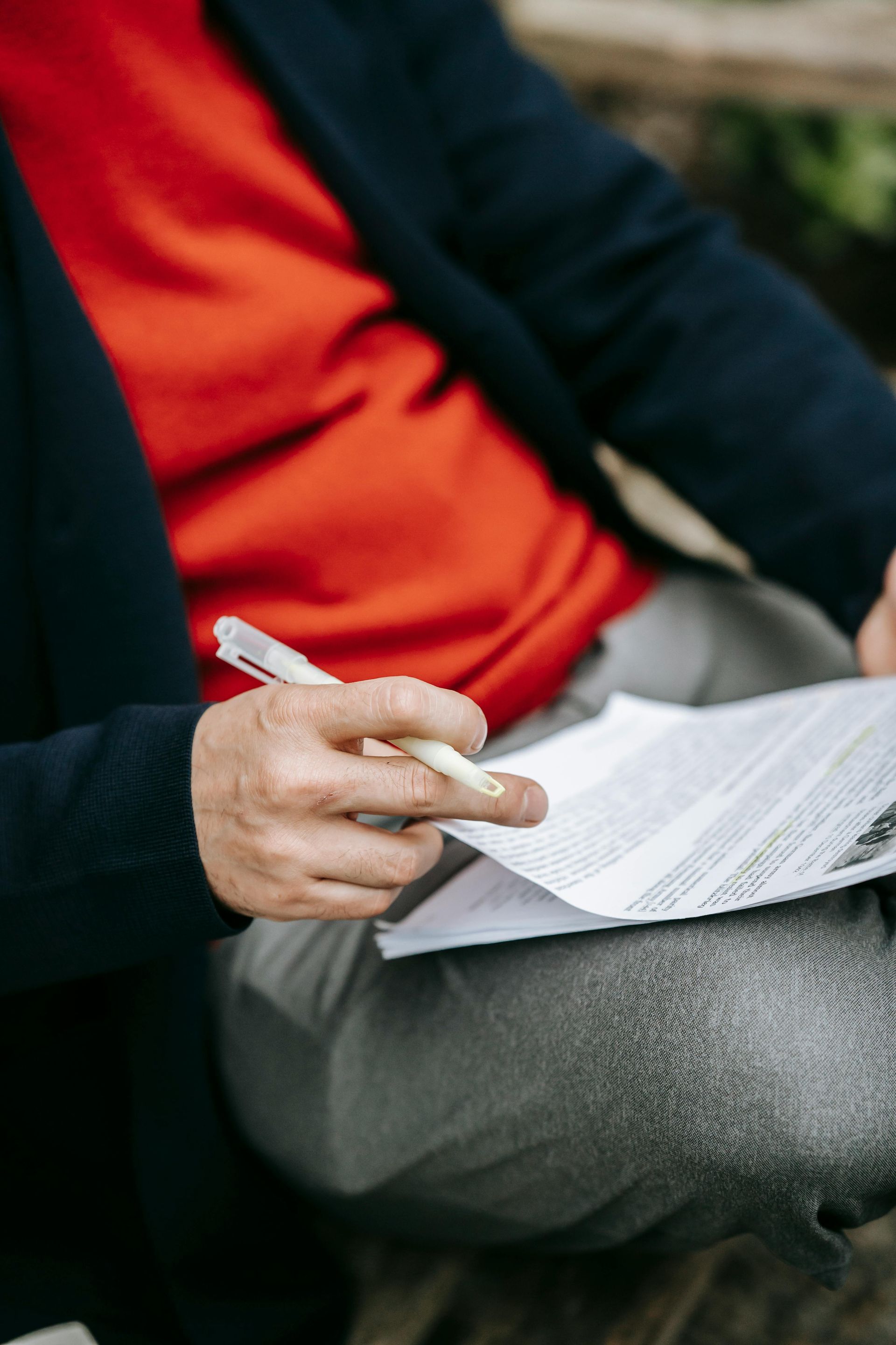 Person holding a pen, writing on papers while sitting. Red shirt and gray pants visible.