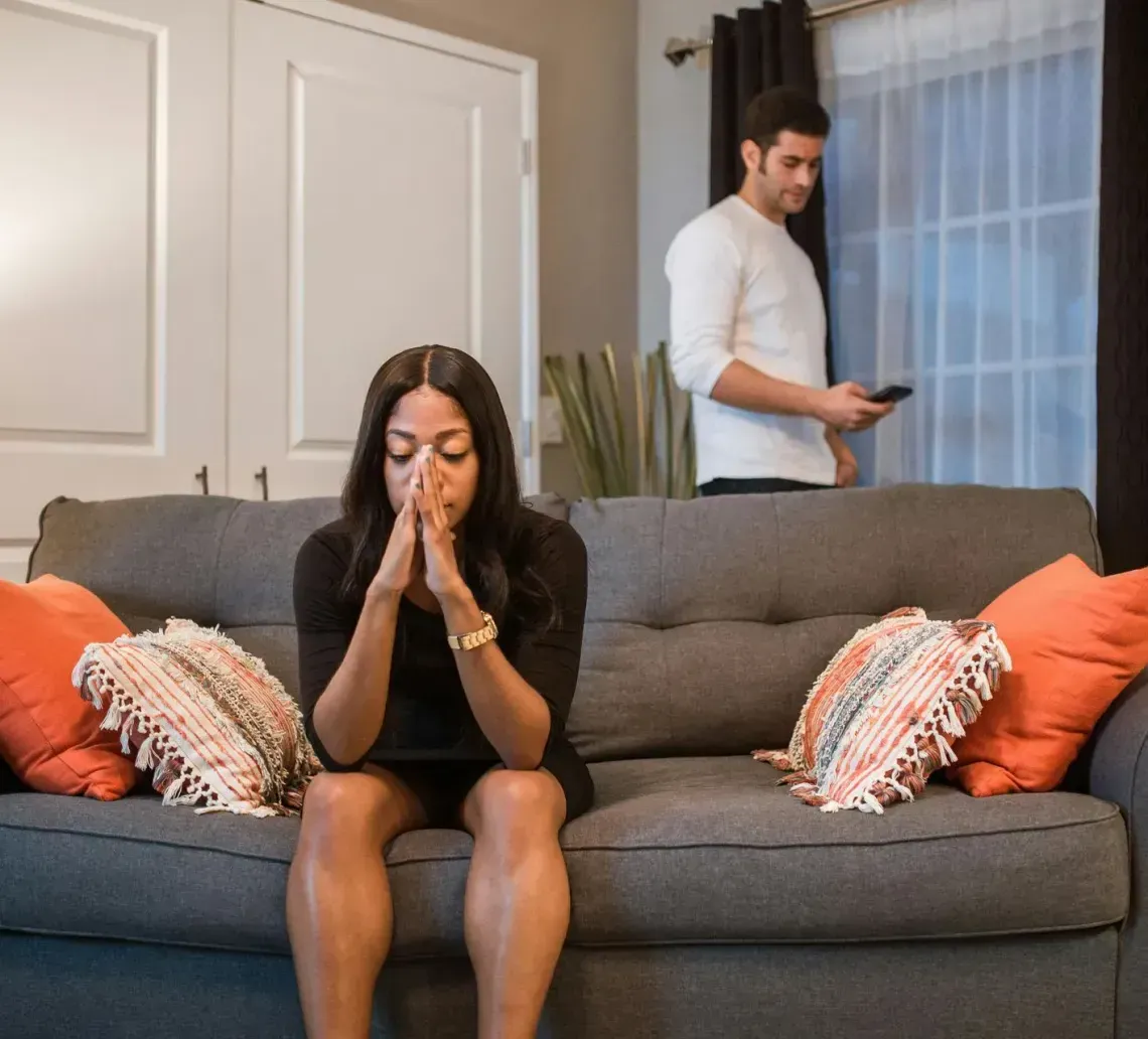 Woman sitting on a gray couch with hands clasped, while a man stands near the window in the background.