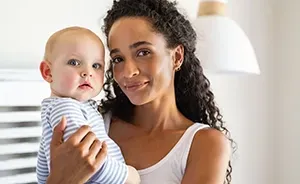 Woman holding a baby in a bright indoor setting, both looking at the camera.