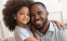 Smiling adult and child sitting close together on a couch, with the child’s arm around the adult.