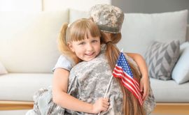 Child hugging a soldier in camouflage, holding a small American flag on a couch