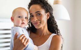 Woman holding a baby indoors, both looking at the camera, with a bright lamp in the background.