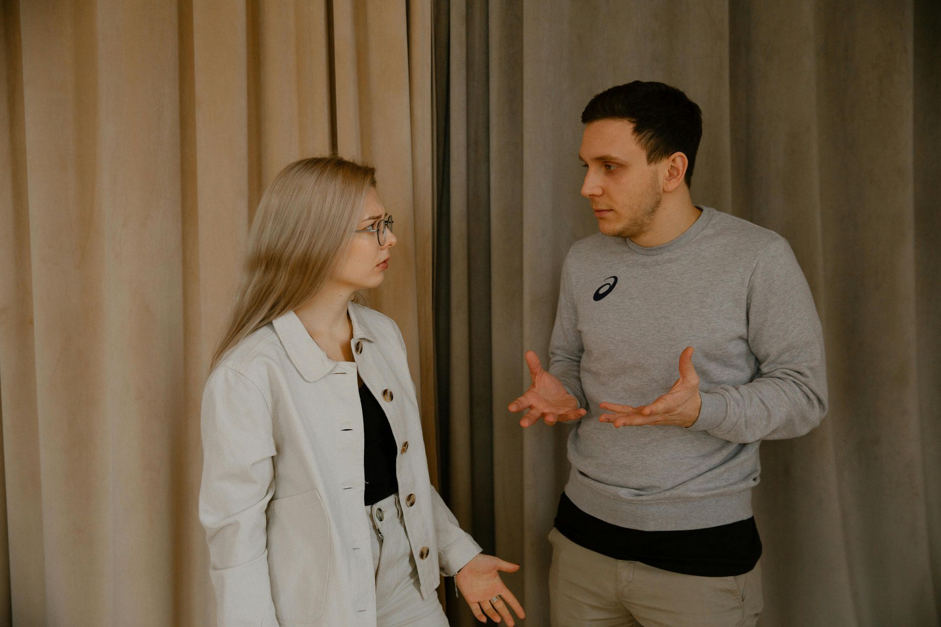 Two people talking in front of beige curtains, one gesturing while the other listens.