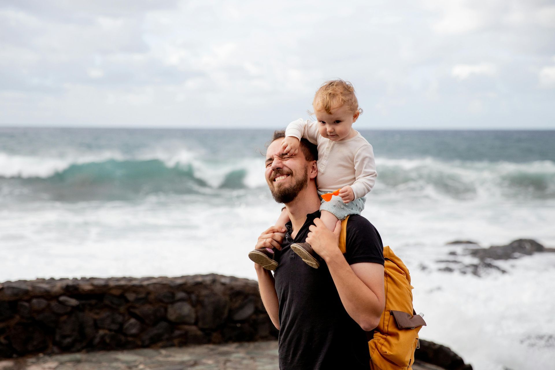 Adult carrying a child on shoulders by the ocean, with waves and rocky shore in the background