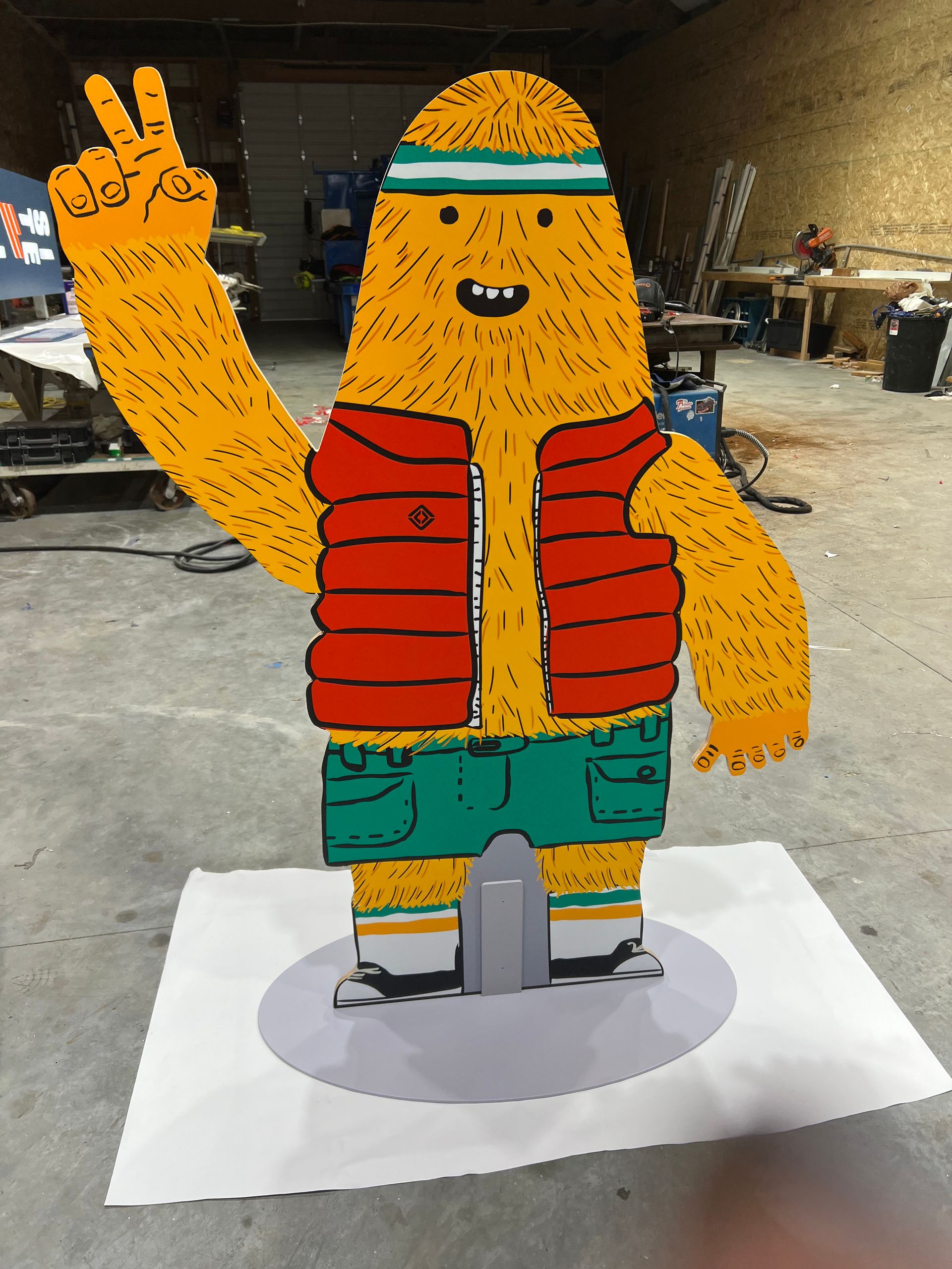 Cartoonish yellow creature in a red vest and green shorts giving a peace sign