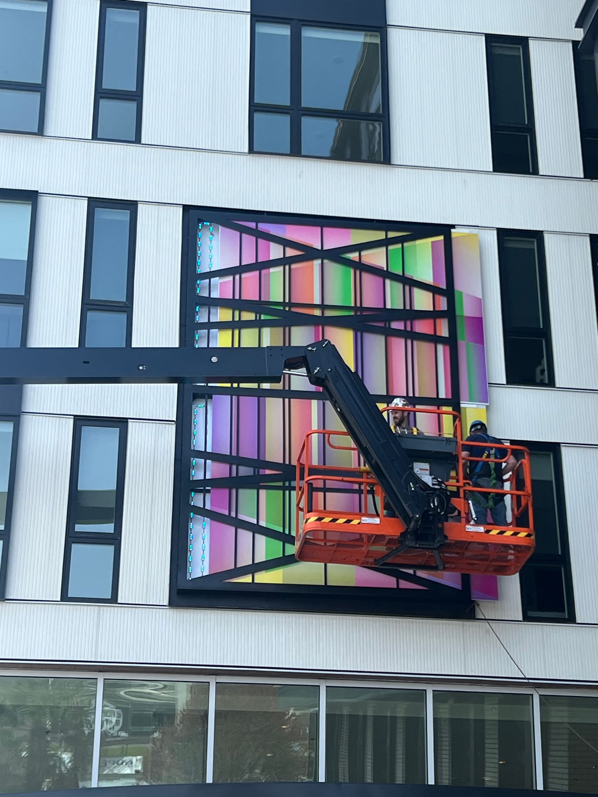 Workers in a lift installing a colorful art piece on a building facade