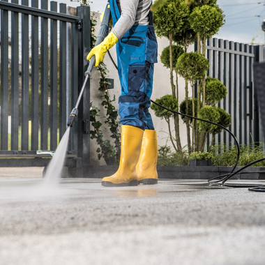 Person power washing a concrete surface with yellow boots and gloves, and blue overalls.