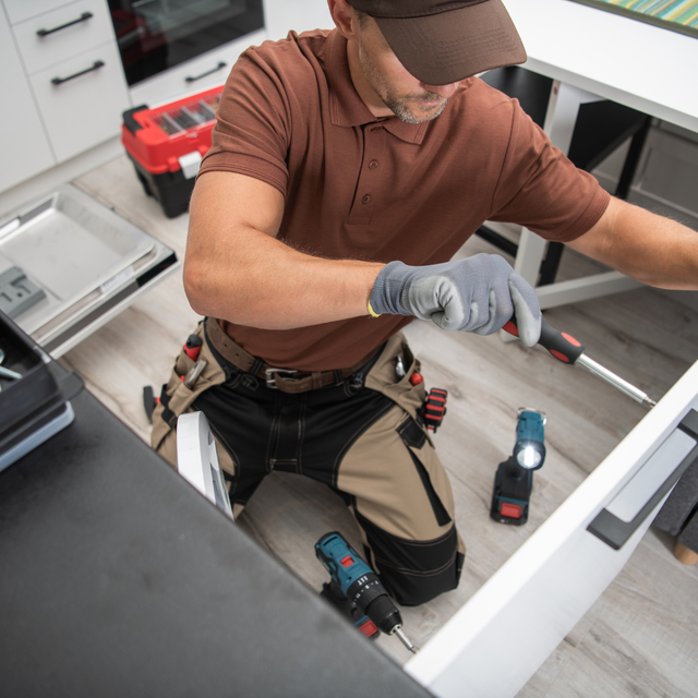 Person kneeling, repairing cabinet, wearing gloves, holding screwdriver, with tools, in kitchen setting.