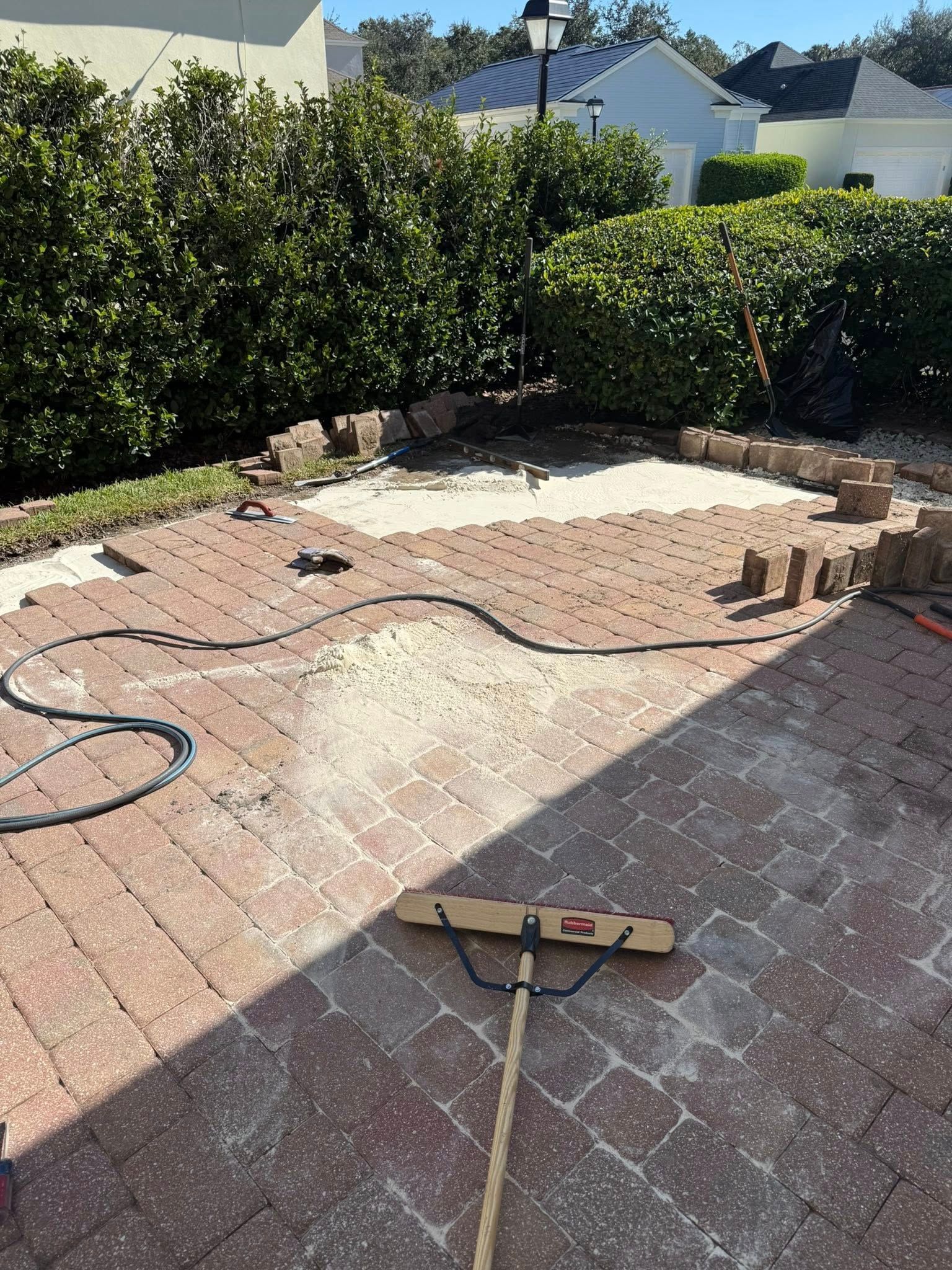 Brick patio being constructed outdoors. Red bricks, a black hose, and a broom are visible.
