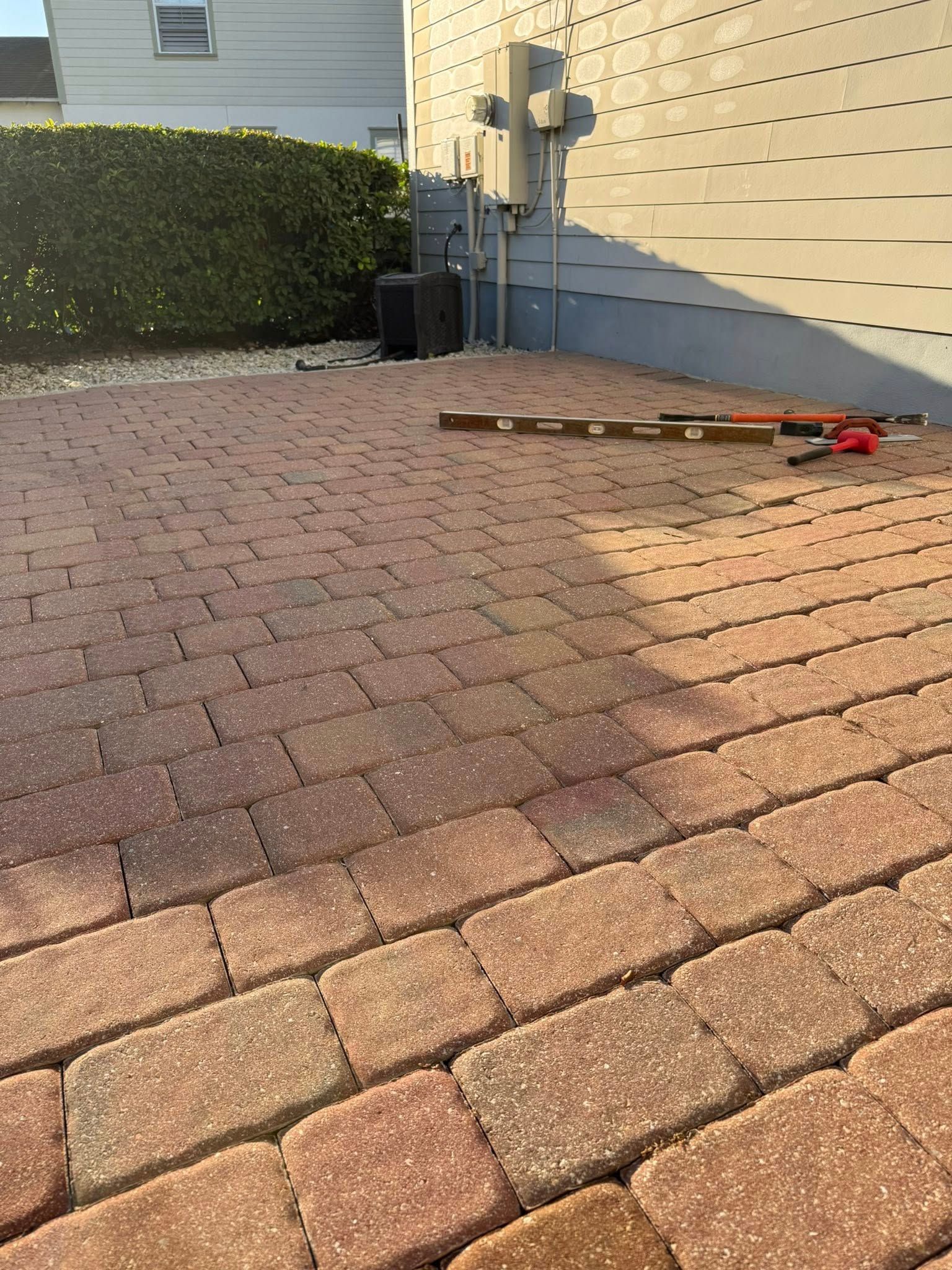 Red brick patio next to a gray wall. Tools and a hedge are visible.